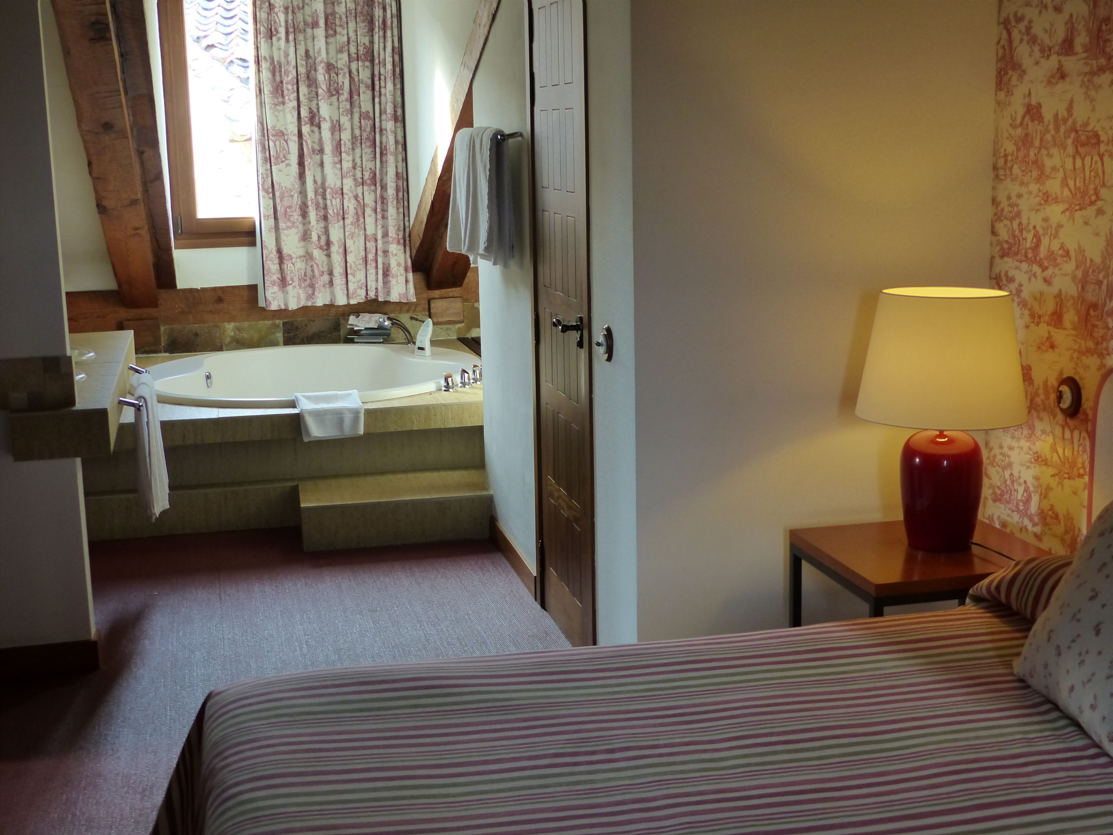 A bedroom with a bed, a nightstand with a lamp, a bathroom with a bathtub, and a window with curtains.