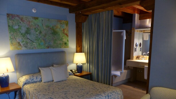 A bedroom with a bed, two bedside tables, a nightstand, a large abstract painting, a bathroom with a sink and mirror, and a chair. - Hostería del Arco (Pedraza)