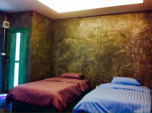 Una habitación con dos camas, una con cubrecama rojo y la otra con cubrecama azul. Hay una puerta verde y una lámpara en el techo.