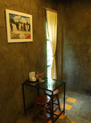A small room with a glass table, two wooden stools, a framed painting of a waterfall, and a window with curtains.