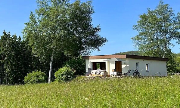 Una casa de un piso con un porche, rodeada de árboles altos y un campo cubierto de hierba.