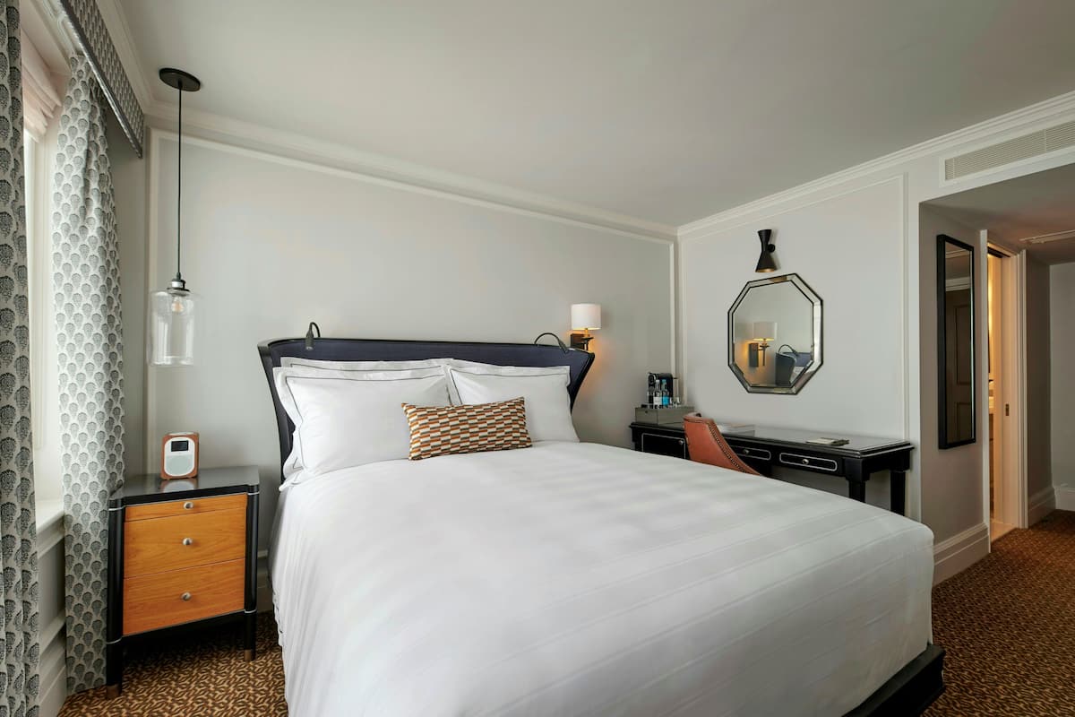 Ein Hotelzimmer mit einem großen Bett, einem Nachttisch, einem Schreibtisch, einem Stuhl, einem Spiegel und gemusterten Vorhängen.