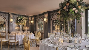 A formal dining room with tables set for a meal, featuring gold chairs, white tablecloths, and floral centerpieces.