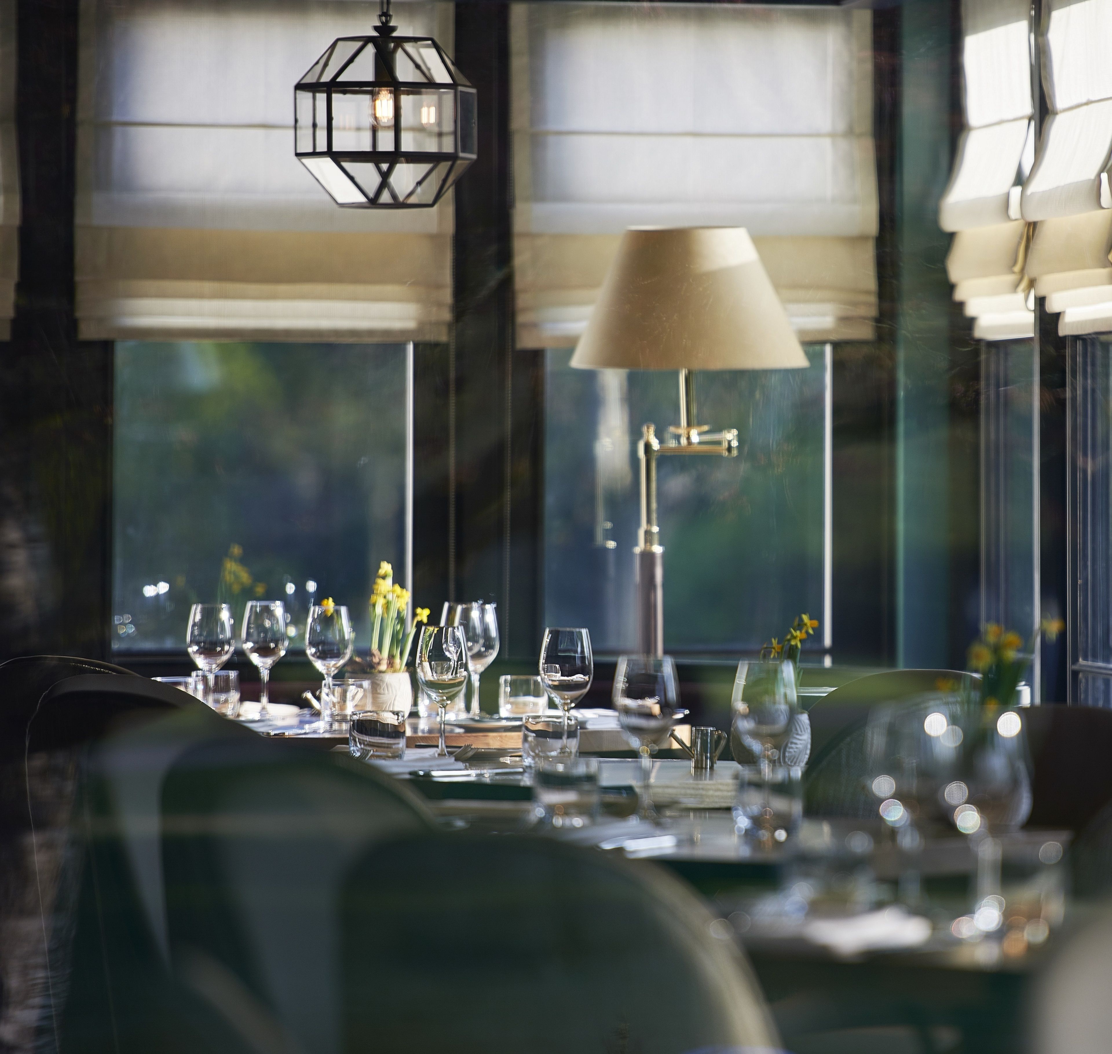 A dining table set with multiple wine glasses and a vase with flowers, with a lamp and hanging light fixture above.