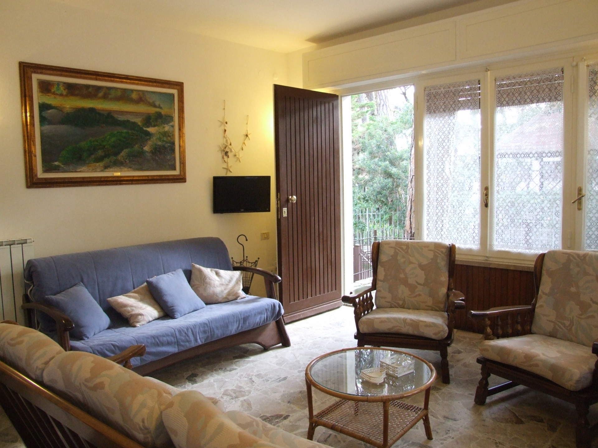 A living room with a blue sofa, patterned armchairs, a glass coffee table, a painting on the wall, and a door leading to an outdoor area.