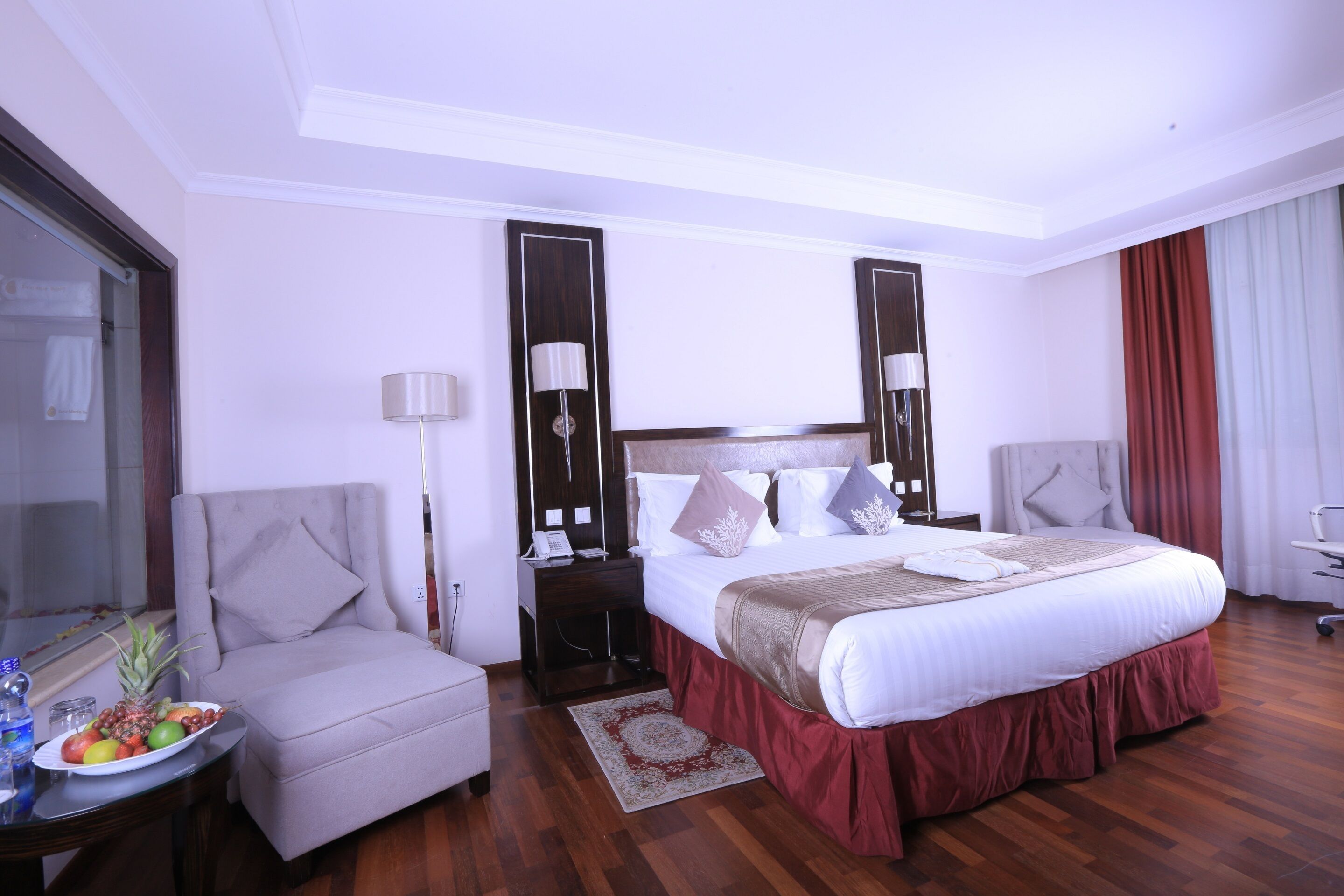 A hotel room with a large bed, a chair, a desk with a lamp, and a small table with fruit.