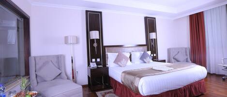 A hotel room with a large bed, a chair, a desk with a lamp, and a small table with fruit.