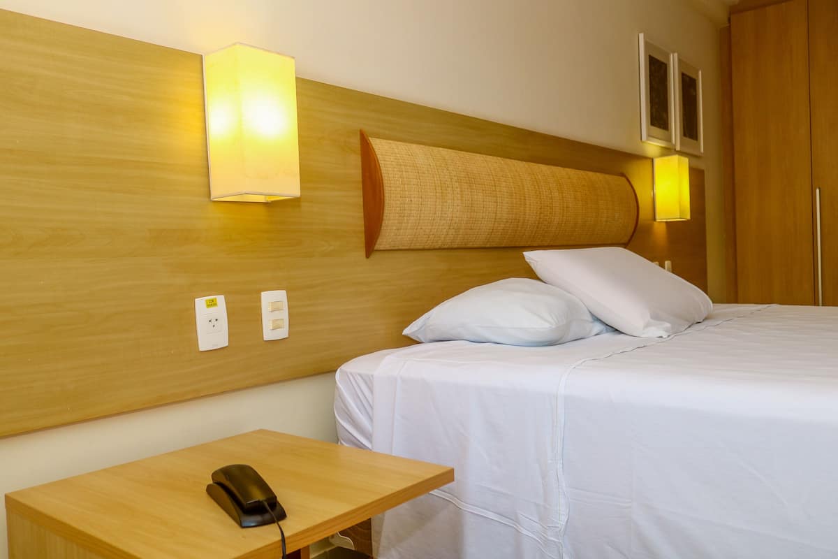 A hotel room with a wooden headboard, a bed with white bedding, a bedside table with a phone, and wall-mounted light fixtures.
