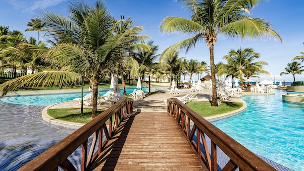 Una terraza de madera que conduce a una piscina rodeada de palmeras y sillas de descanso.