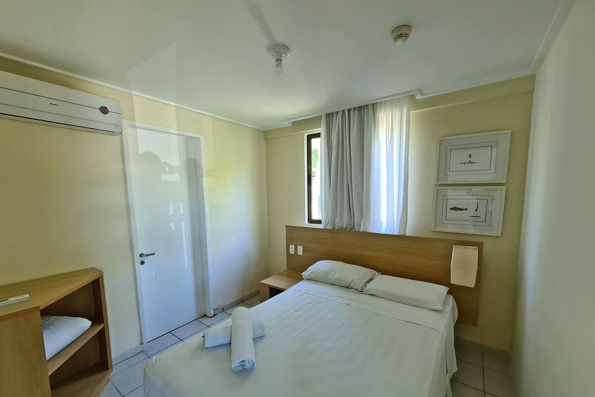 A hotel room with a bed, a wooden nightstand, a desk with a chair, a wardrobe, and a window with curtains.
