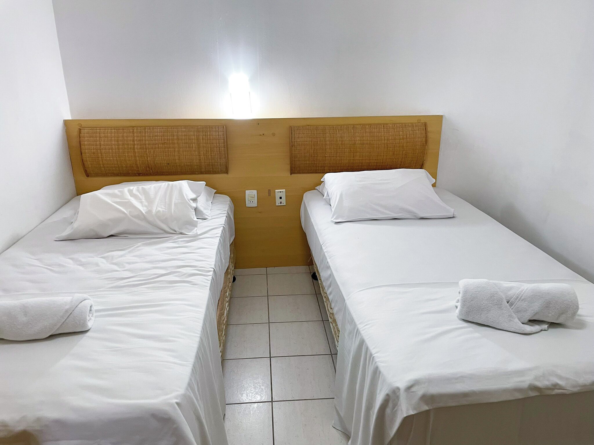 A twin room with two single beds, each with white linens and pillows, a headboard with a woven design, and a small nightstand with a lamp.