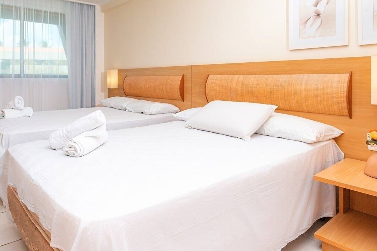A hotel room with two beds, a wooden headboard, white linens, a bedside table with a lamp, and a window with curtains.