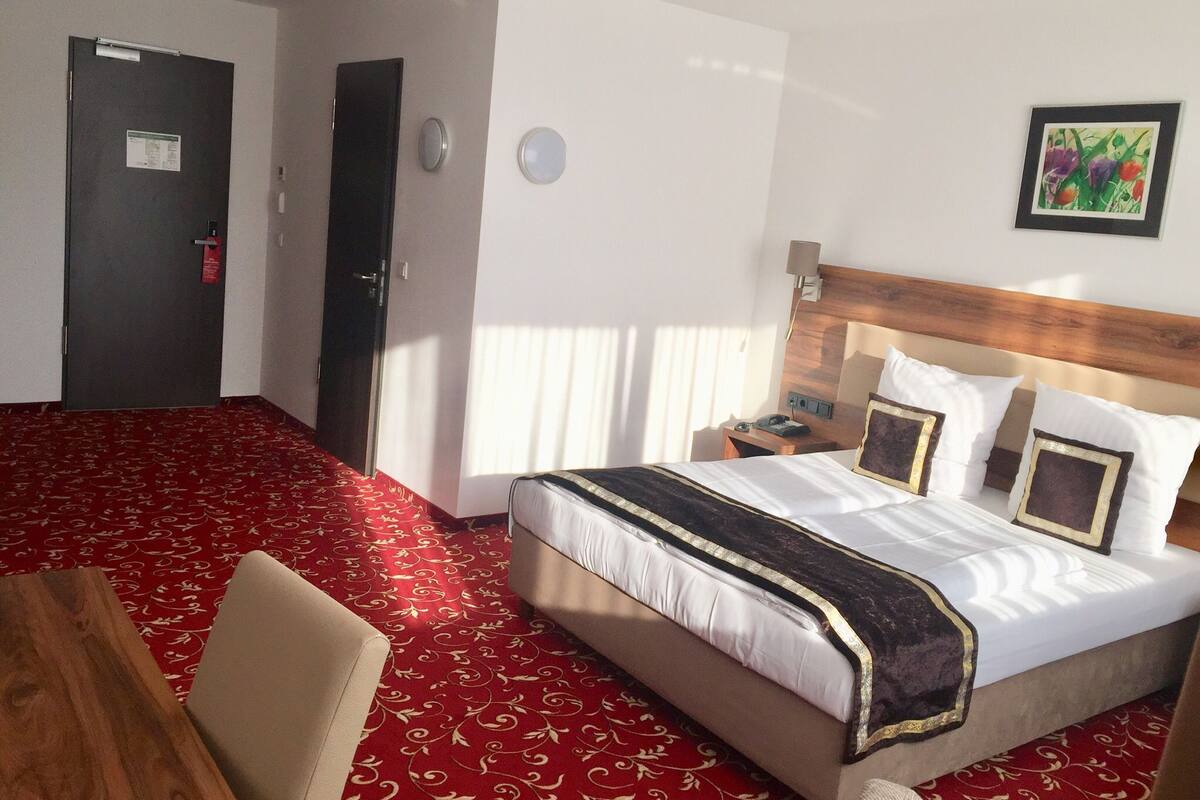 Une chambre d’hôtel avec un grand lit, une tête de lit en bois, une moquette rouge à motifs, une chaise en bois et une porte avec une pancarte.