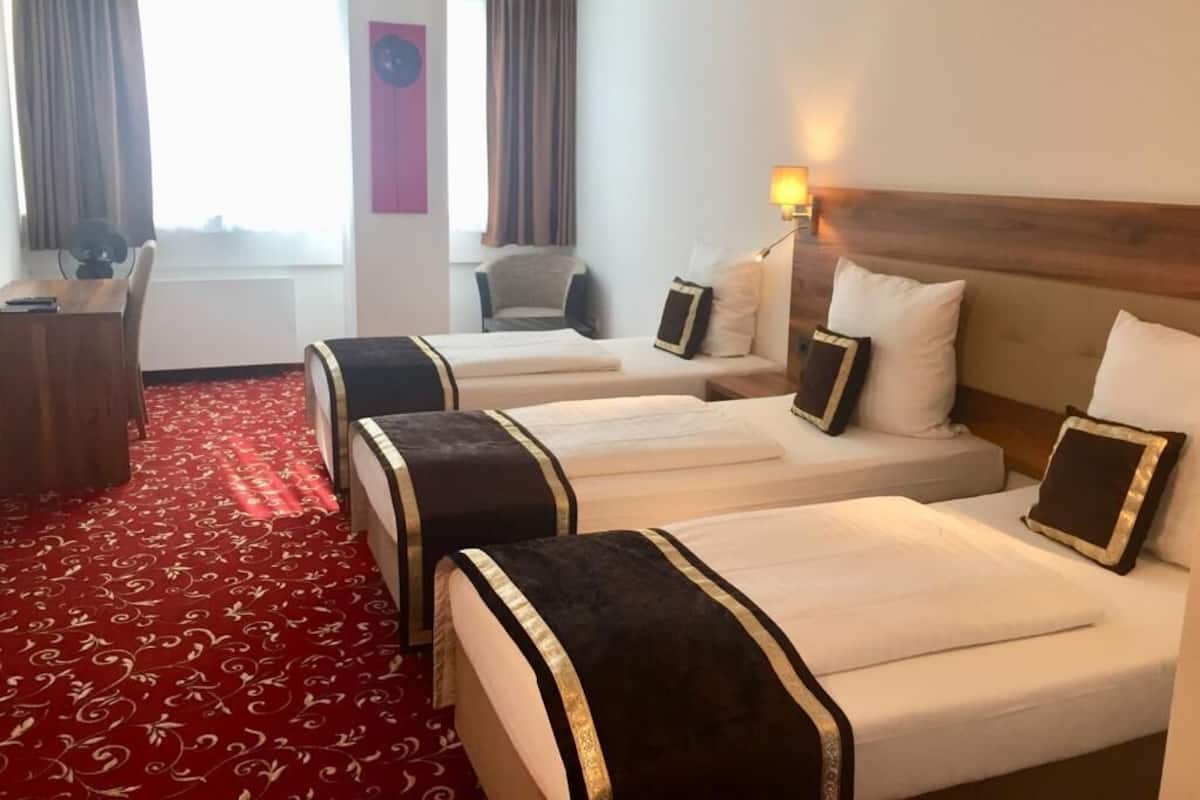 Une chambre d’hôtel avec trois lits, un bureau, une chaise et une moquette rouge.