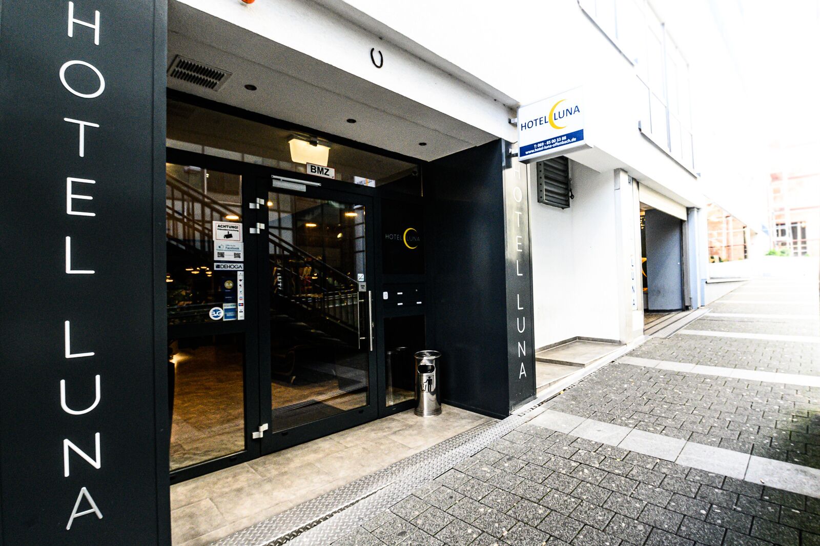 Entrada de um hotel com porta de vidro, placa indicando "HOTEL LUNA" e escadas que levam ao interior.