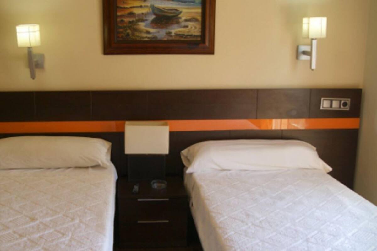 A hotel room with two single beds, a nightstand, a painting on the wall, and two wall-mounted lamps.