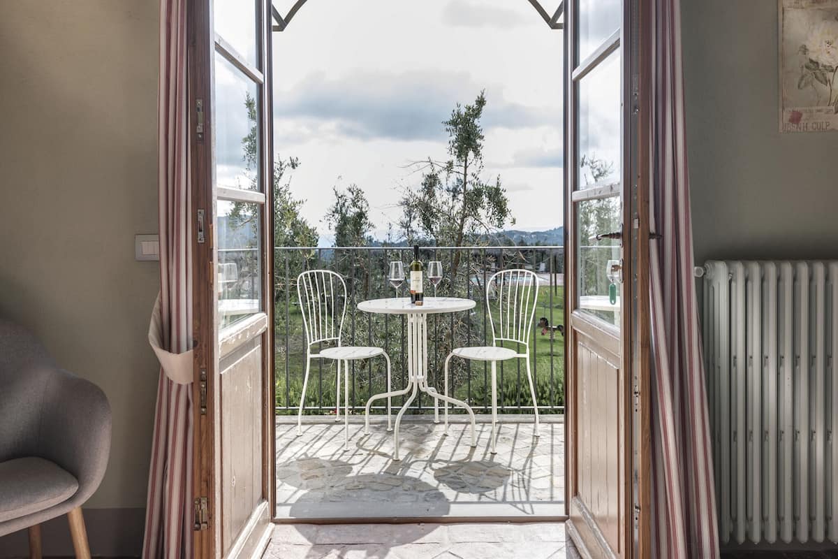 Une porte ouverte donne sur un balcon où se trouvent deux chaises blanches et une table, ainsi qu’un radiateur et un tableau accroché au mur.