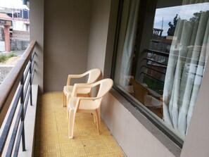 A balcony with two plastic chairs, a tiled floor, and a view of buildings through a window with a curtain. - Raahat Inn Kodaikanal (Kodaikanal)