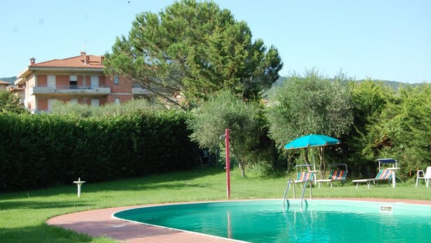 A swimming pool with a red diving board, surrounded by a grassy area and a hedge. There are lounge chairs and a blue umbrella.