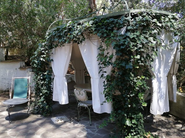 A covered outdoor seating area with white curtains, a table, and chairs, surrounded by greenery.
