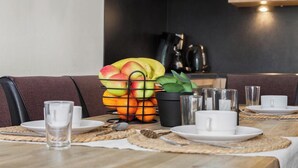 Un comedor con una mesa de madera dispuesta para dos personas, con platos, vasos y cubiertos. Hay una cesta de frutas con manzanas, plátanos y naranjas sobre la mesa, y una planta en maceta cerca.