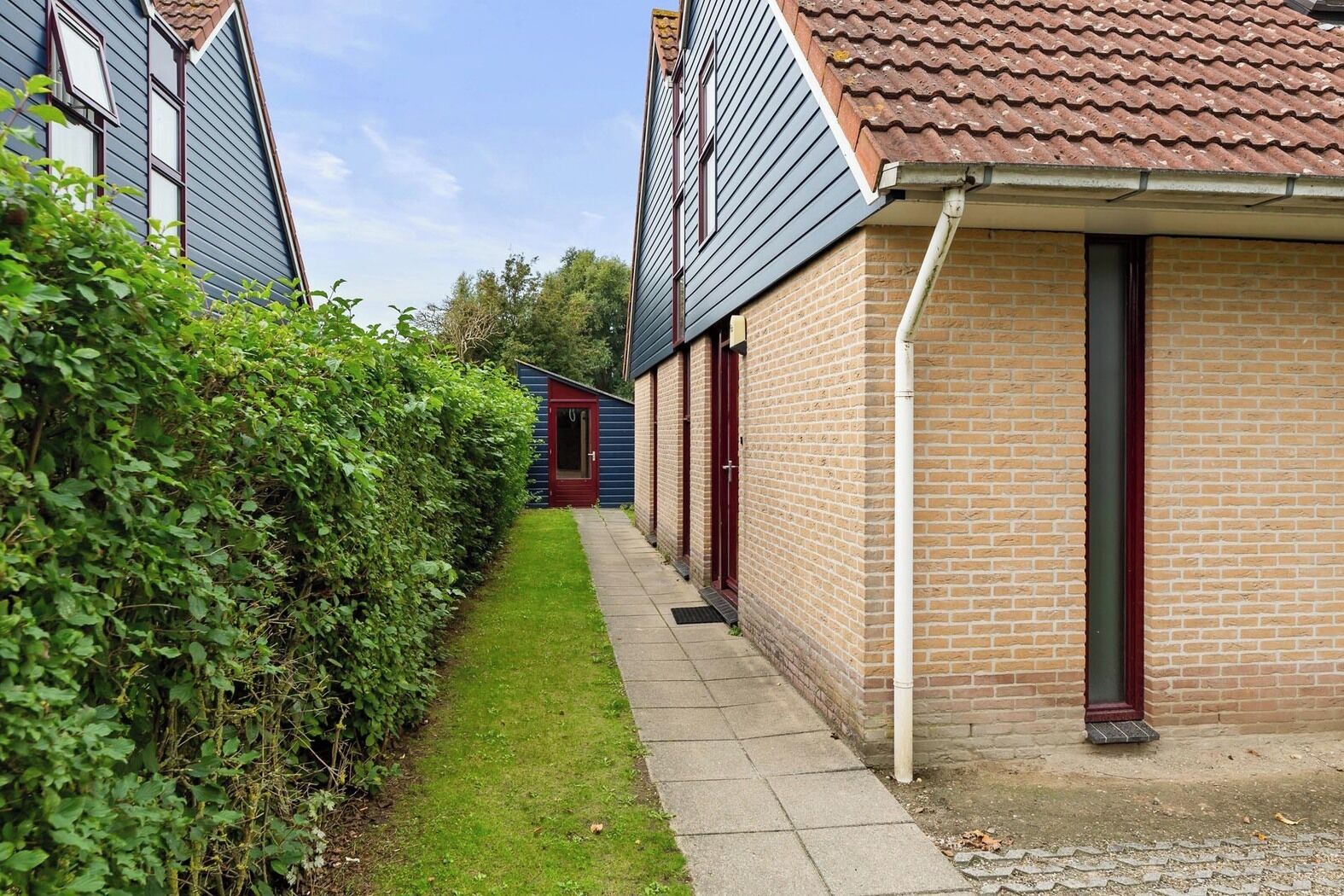 Een smalle pad tussen twee bakstenen huizen, met een kleine tuin en een rode deur.