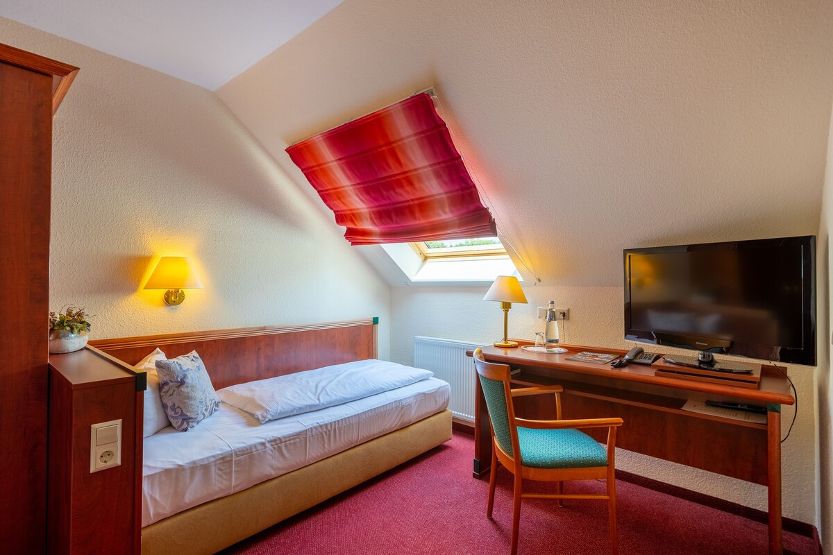 Une chambre d’hôtel avec un lit, un bureau, une chaise, une télévision et une fenêtre avec un store rouge.