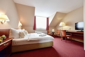 Habitación de hotel con cama, escritorio, televisor y ventana con cortinas.