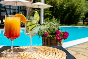 Dos bebidas sobre una mesa trenzada junto a una piscina, con una cesta de mimbre y flores al fondo.