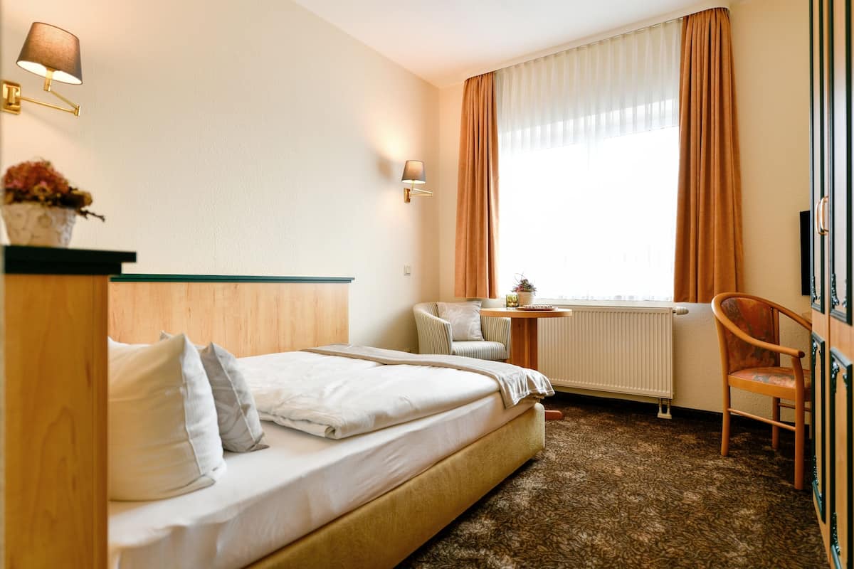Une chambre d’hôtel avec un lit, une chaise, une petite table, une lampe et une fenêtre avec des rideaux.