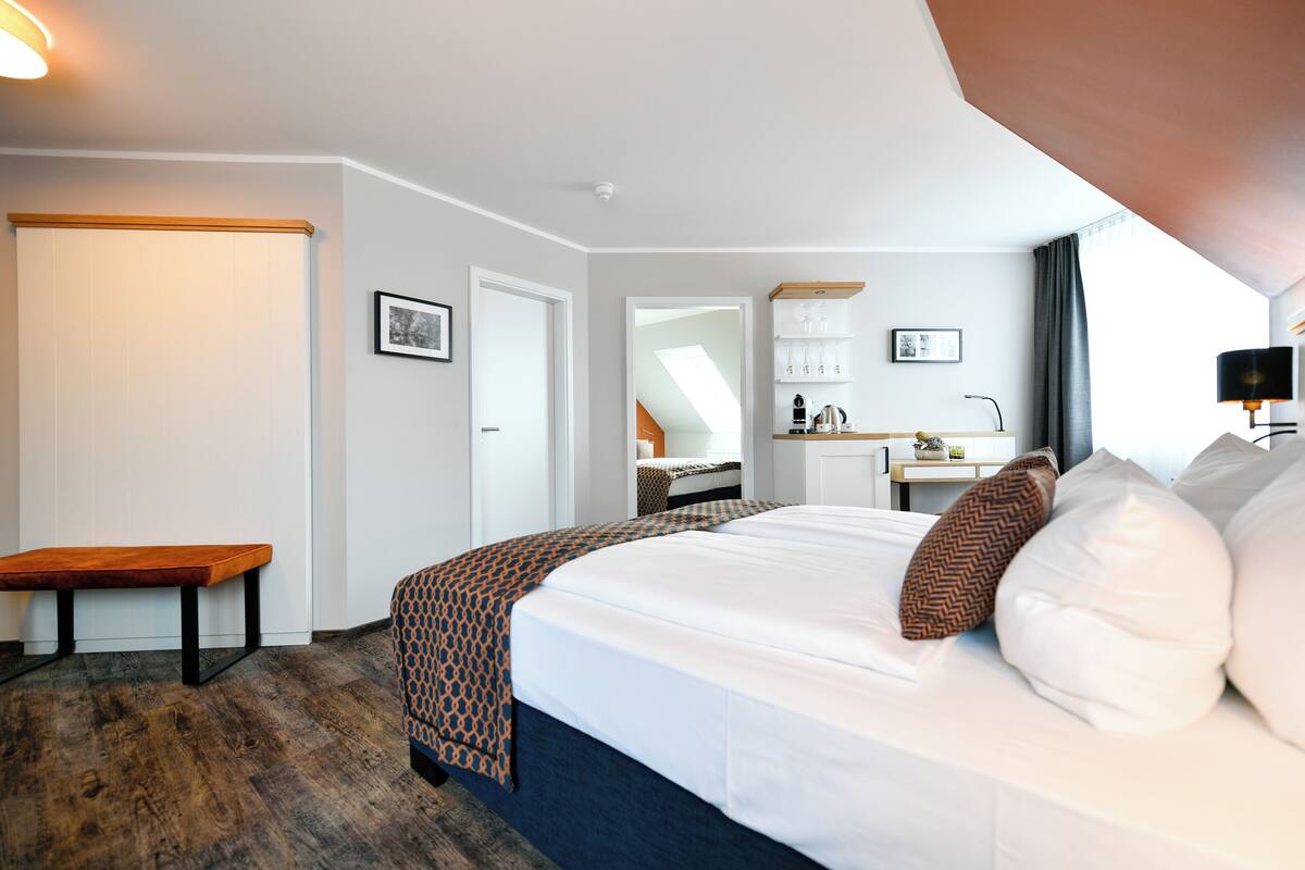 Une chambre d’hôtel avec un lit, une banquette, un bureau et une salle de bain visible à travers une porte ouverte.