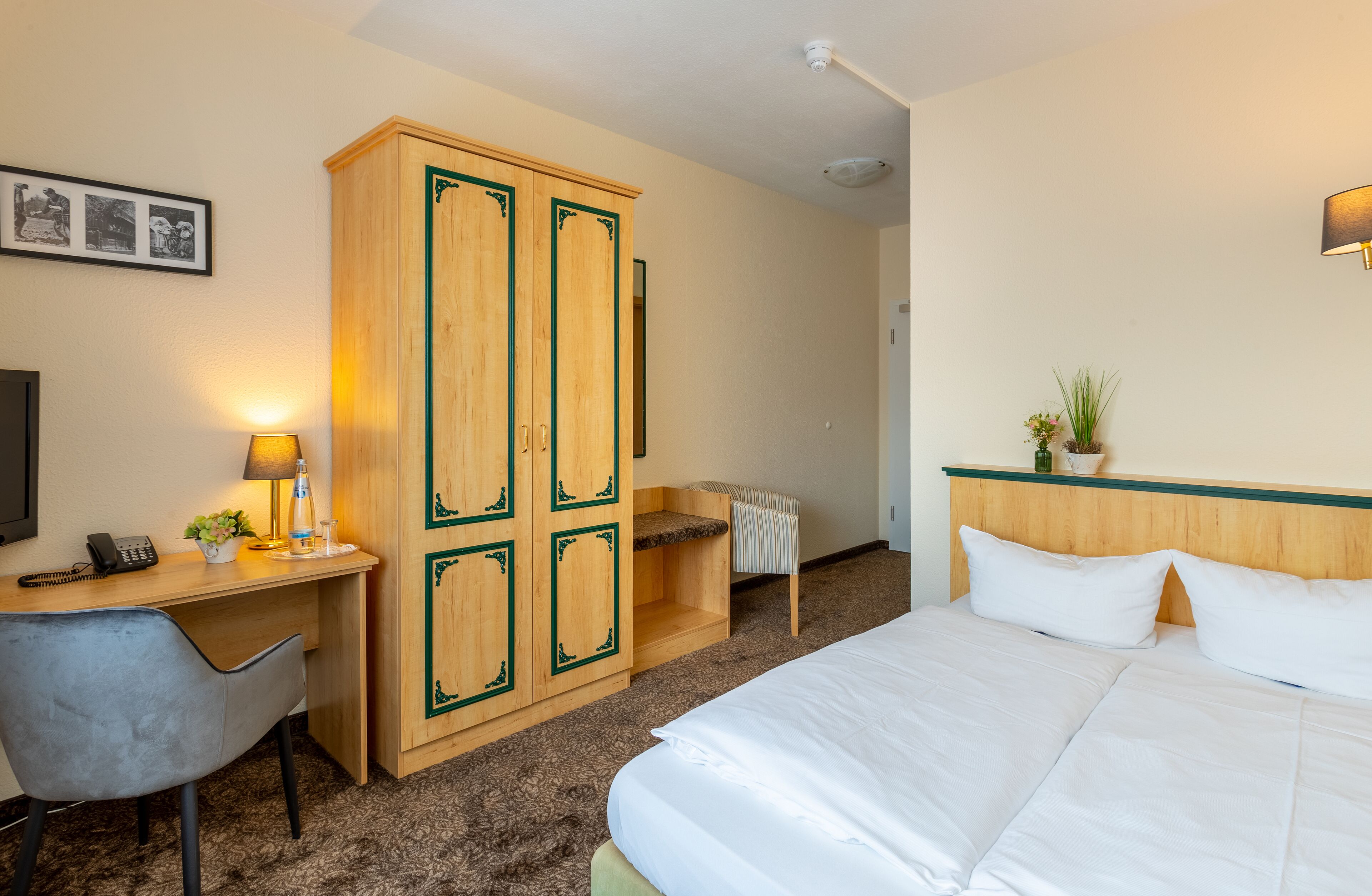 Una habitación de hotel con una cama, un escritorio con lámpara, un armario y una planta pequeña en la mesita de noche.