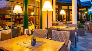 An outdoor seating area with wicker furniture and lamps, a green building in the background, and a view of the interior through large windows.