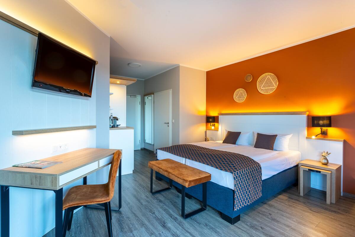 Une chambre d’hôtel moderne équipée d’un lit, d’une table de chevet, d’un banc, d’un bureau, d’une chaise et d’un téléviseur fixé au mur.