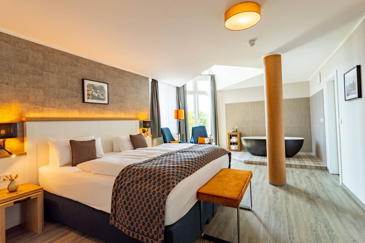 Une chambre d’hôtel moderne dotée d’un grand lit, d’un coin salon avec un banc, d’une grande fenêtre et d’une baignoire.