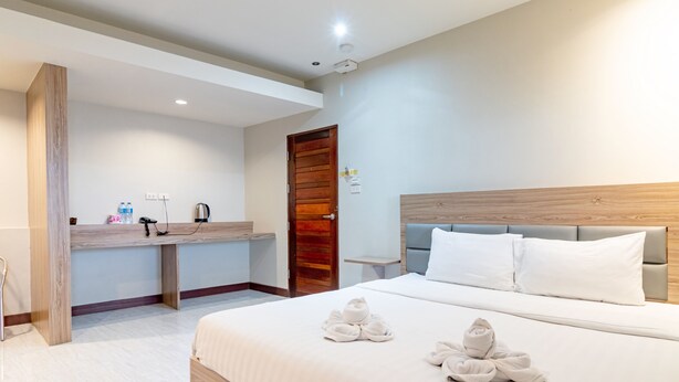 A hotel room with a wooden headboard, a large bed with white bedding, a wooden bench at the foot of the bed, and a small table with a kettle and bottles. - Oun Residence (Hat Yai)