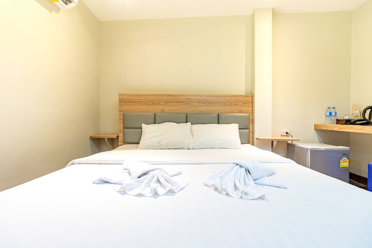 A hotel room with a bed, a wooden headboard, a nightstand, a mini-fridge, and a shelf with toiletries.