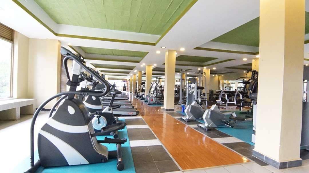 A gym with various exercise equipment, including treadmills and weight machines, arranged in a spacious layout with a tiled floor and green ceiling accents.