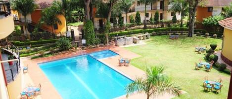 Piscina con sillas de descanso y mesas, rodeada de césped cuidado y árboles.