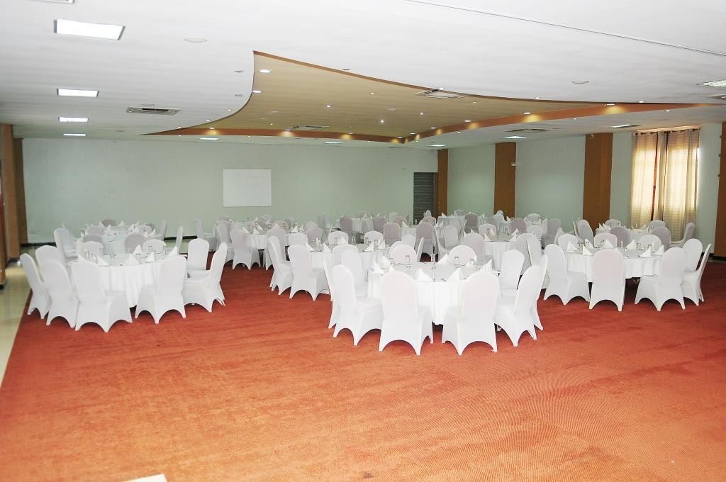 Une grande salle de conférence avec de longues rangées de tables et de chaises blanches, un sol recouvert de moquette et un tableau blanc accroché au mur.