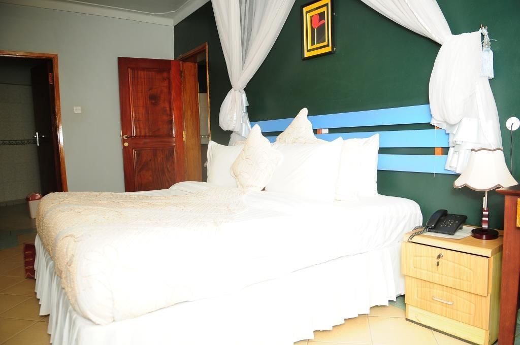 Una cama bien hecha con sábanas blancas, una mesita de noche de madera con un teléfono, y una pared verde con un cuadro enmarcado.