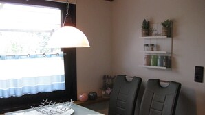 Un comedor con una mesa lista para dos personas, una lámpara colgante y una estantería con utensilios de cocina.