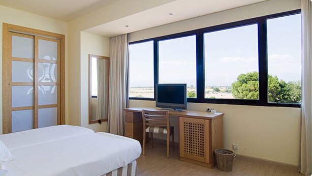 Habitación de hotel con cama, escritorio con televisor, ventana con vista y puerta con paneles de vidrio esmerilado.