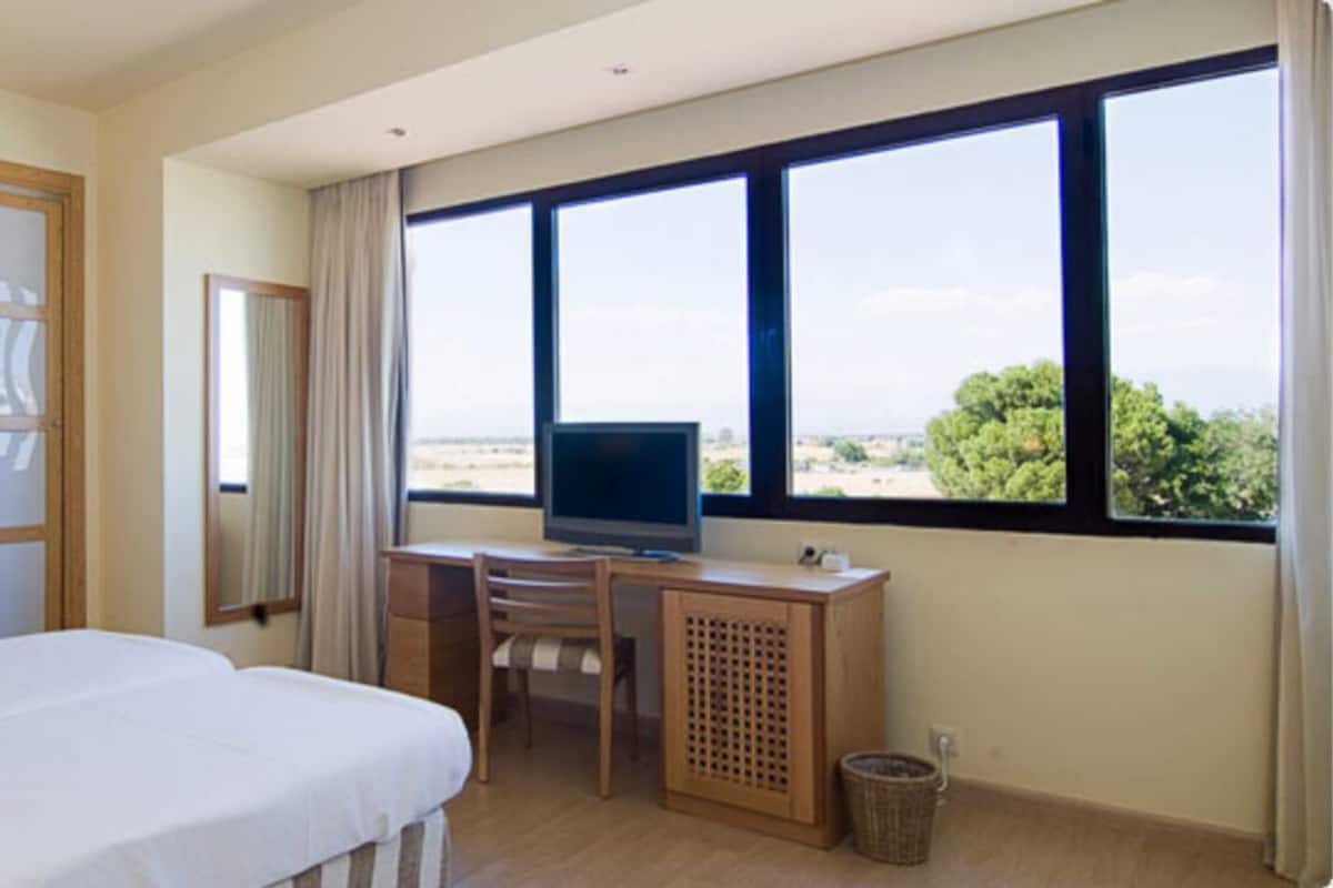 A hotel room with a bed, a desk with a television, a window with a view, and a door with frosted glass panels.