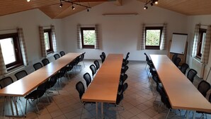 Un salón de conferencias con largas mesas y sillas, vigas de madera en el techo y ventanas con cortinas.