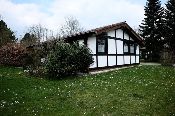 Una casa tradicional de entramado de madera con tejado inclinado, rodeada de un césped cuidado y plantas con flores.