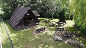 Una cabaña de madera con techo inclinado, rodeada de un área verde con mesas de picnic y un brasero.