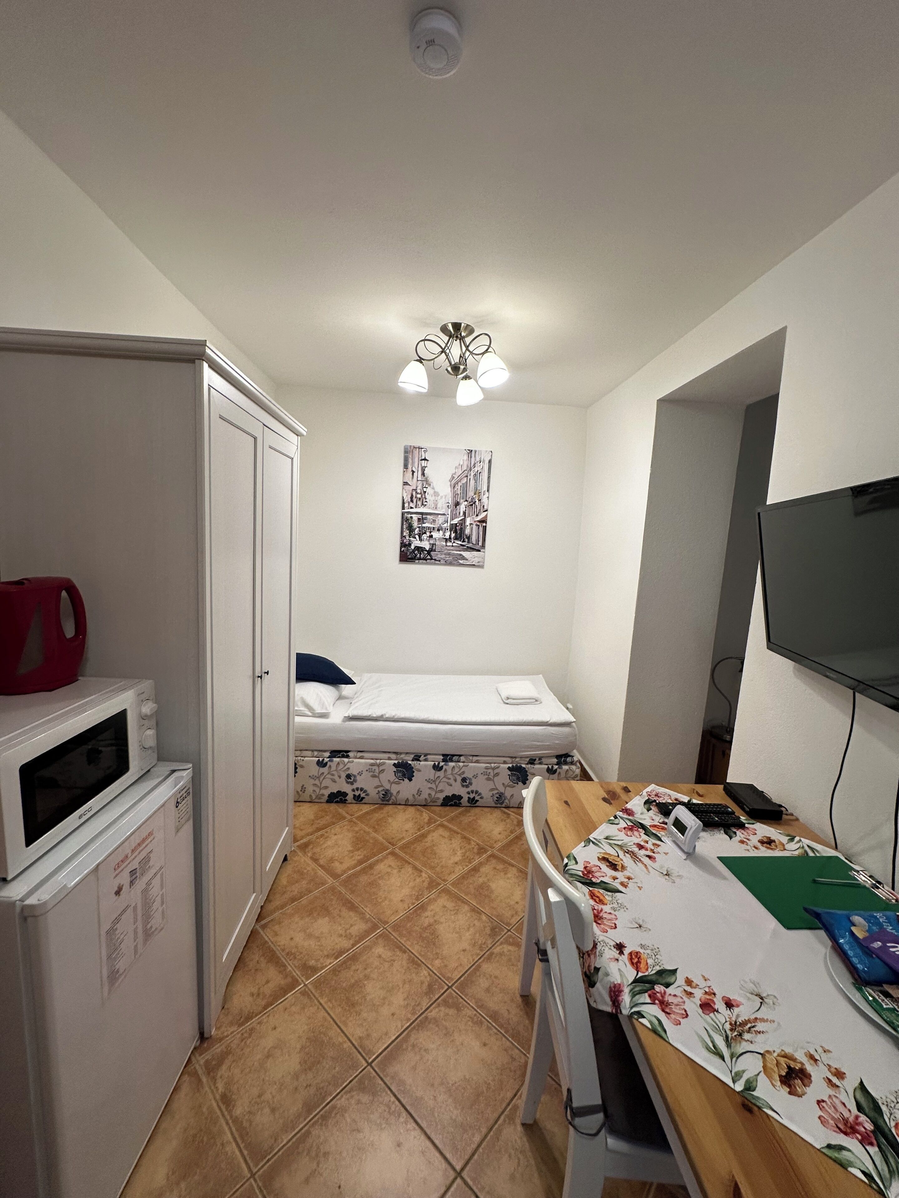 A compact room with a bed, a kitchenette, a small table with a chair, a ceiling light, and a wall-mounted TV.