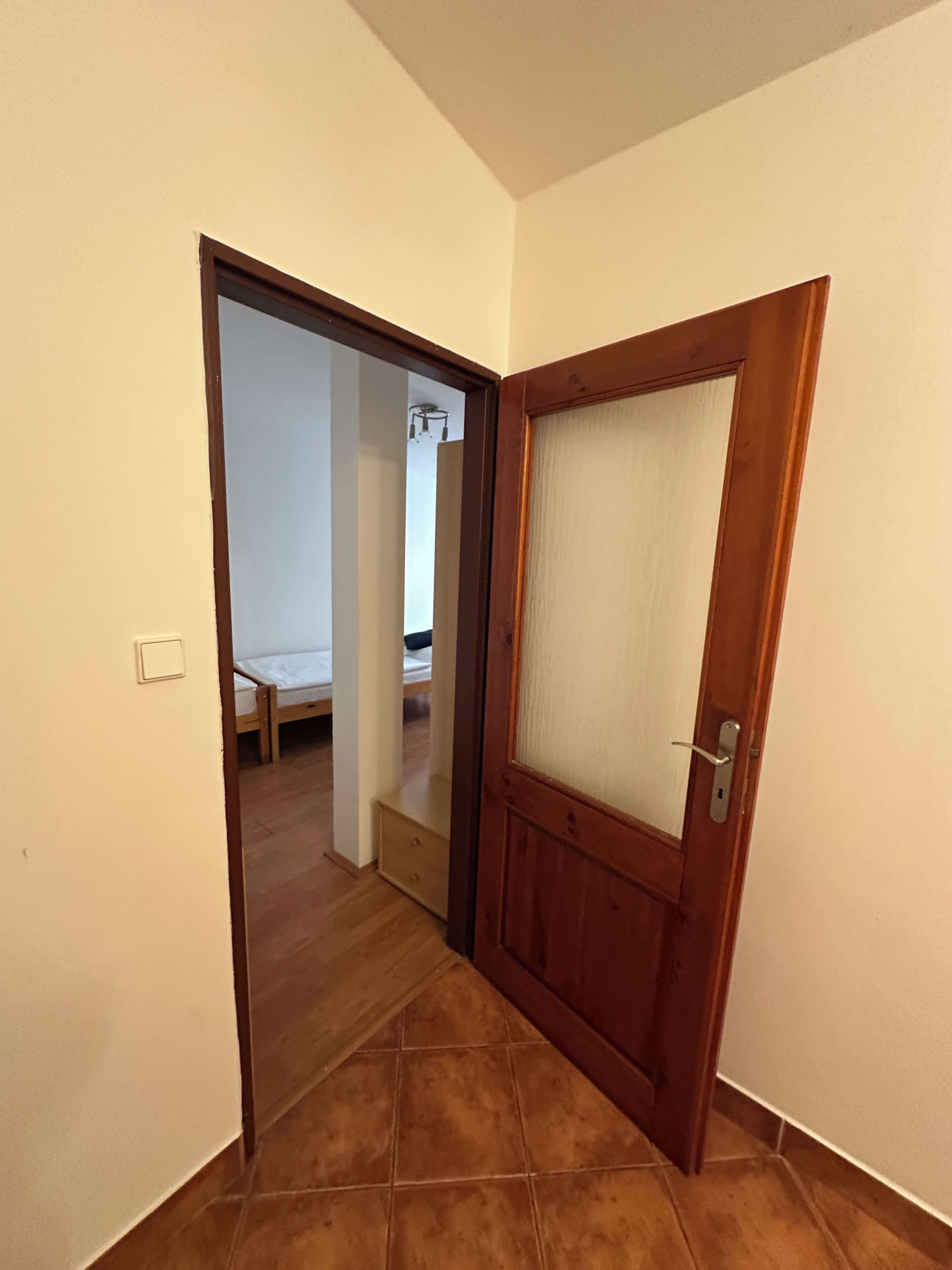 Un pasillo con una puerta de madera, piso de baldosas y una cama en la habitación contigua.