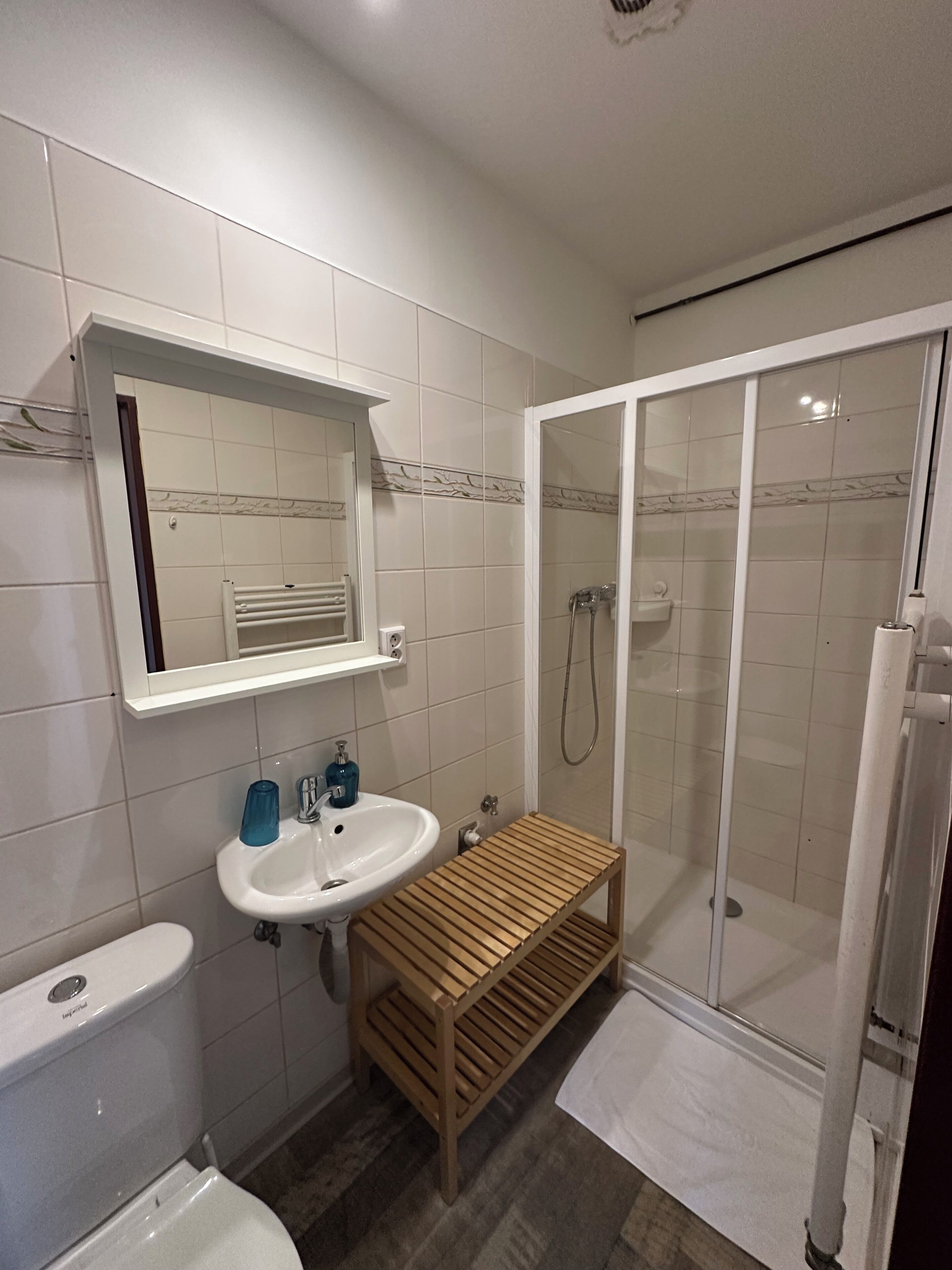 A bathroom with a toilet, sink, mirror, shower, and a wooden shelf.
